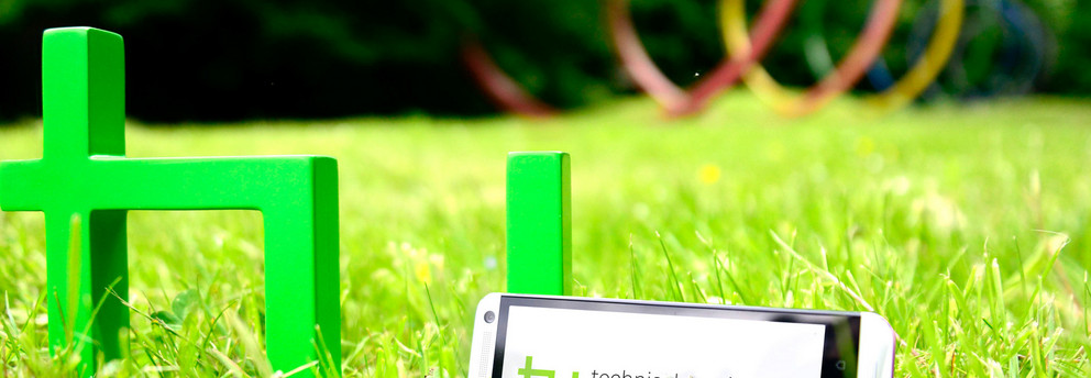 TU-App Ein grünes TU-Logo steht auf grünem Rasen. Daran lehnt ein Smartphone.